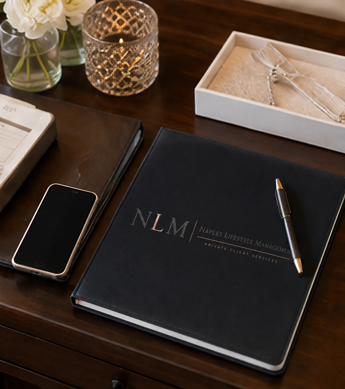 Black leather folio displaying the Naples Lifestyle Management logo on an elegant desk with a phone, pen, flowers and candle